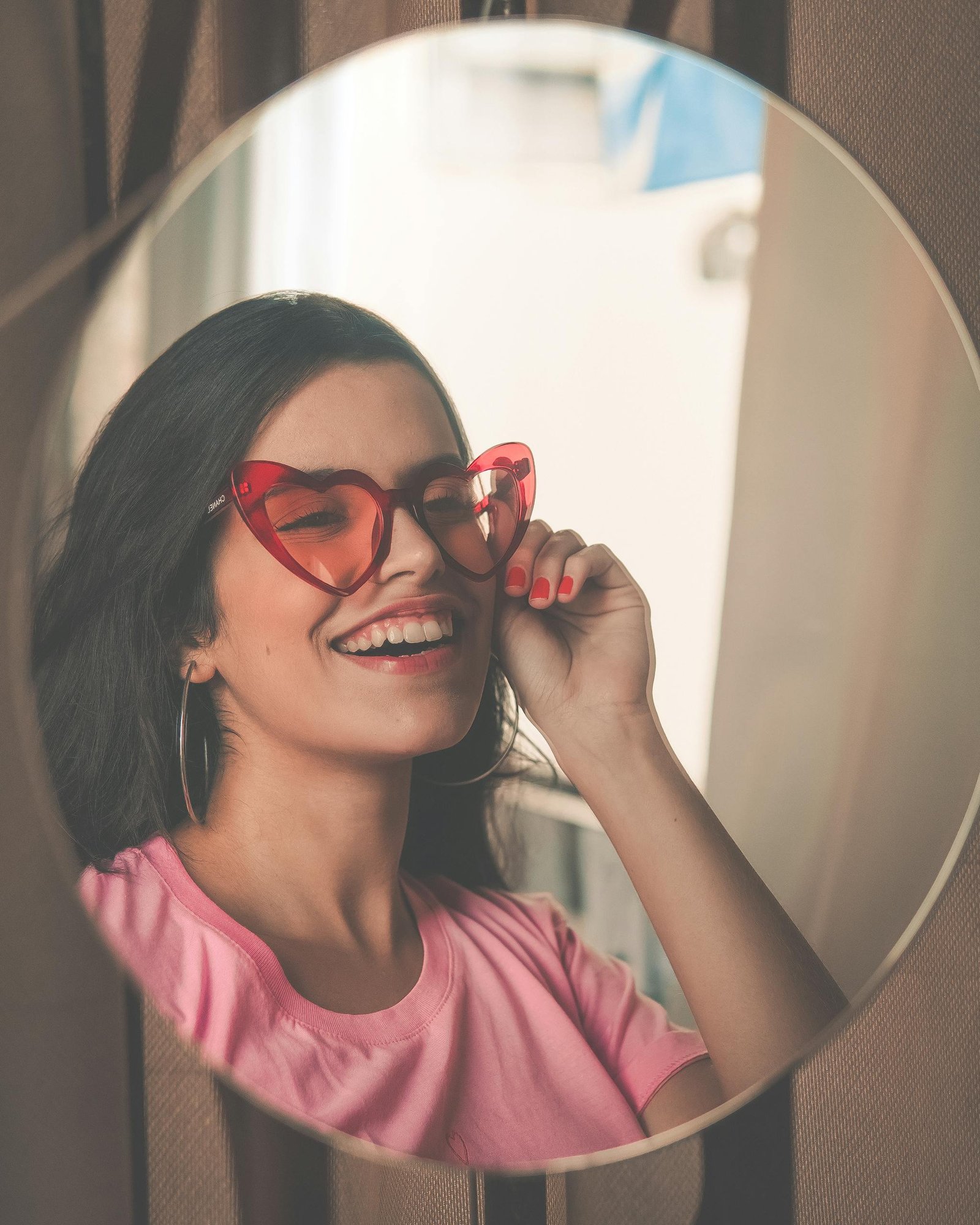 Stylish woman wearing heart-shaped sunglasses smiles joyfully at her reflection in a mirror.